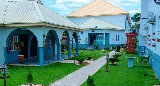 The House 7 compound at midday, a blue arcaded building along a lawn, with a paved path and lamp posts.