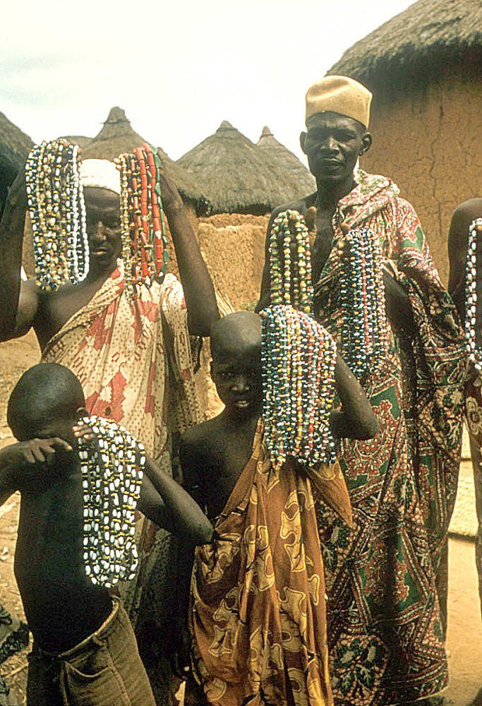 Nupe people holding up long strands of traditional glass beads outside a thatched compound in Bida.