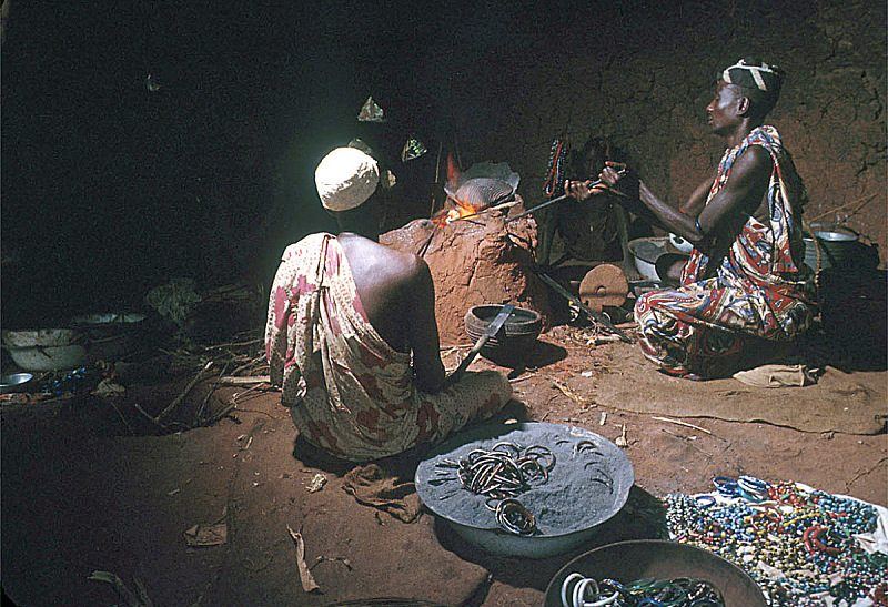 Two people seated on the ground working a low fire, with trays of beads beside them.