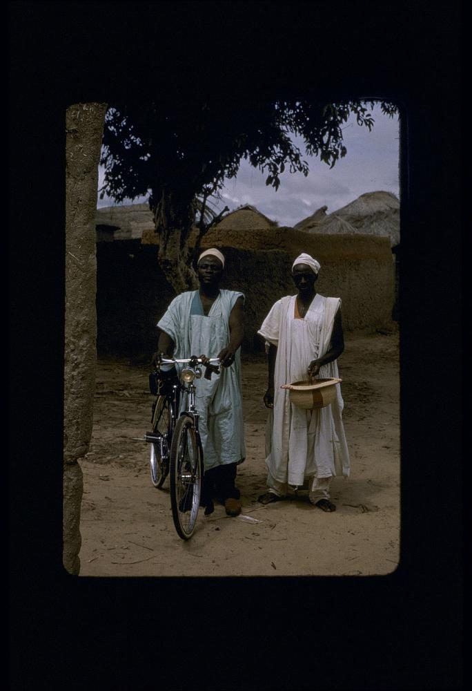 Two men in embroidered gowns standing with a bicycle outside a mud-walled compound.