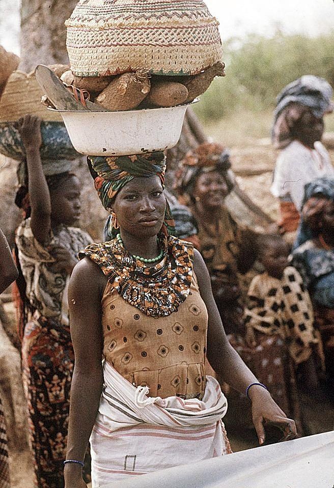 A woman in a beadwork collar and printed wrapper carrying a cane basket on her head in a crowd.
